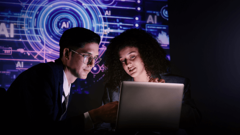 Two professionals collaborate on a laptop with AI graphics displayed on digital screens in the background.