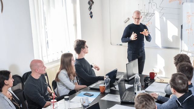 Business professional presenting to colleagues during a technology strategy meeting with laptops and a whiteboard.