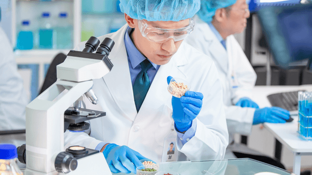 Laboratory technician in white coat examining sample under microscope with blue gloves