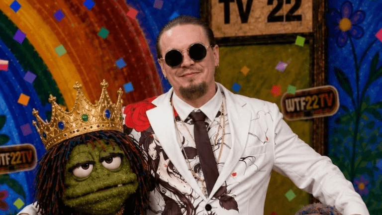 A man in a flamboyant suit poses with a crowned puppet against a colorful backdrop