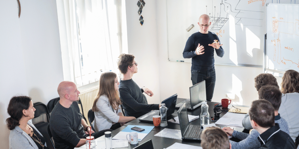 Investors Turn Attention to Big Tech Earnings as AI Spending Faces Its Next Major Test Business professional presenting to colleagues during a technology strategy meeting with laptops and a whiteboard.