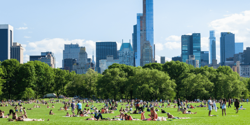 Crowds relax and walk across a large green urban park surrounded by trees and city skyscrapers.